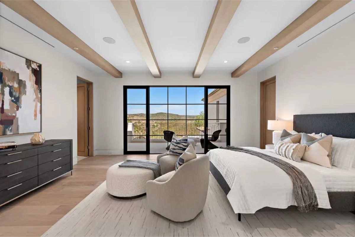 Cozy bedroom with exposed wooden beams, a large glass door leading to a scenic balcony, and neutral-toned furnishings. 