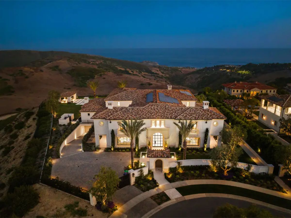 Mediterranean-style mansion stands atop rolling hills with panoramic views of the ocean at dusk. 