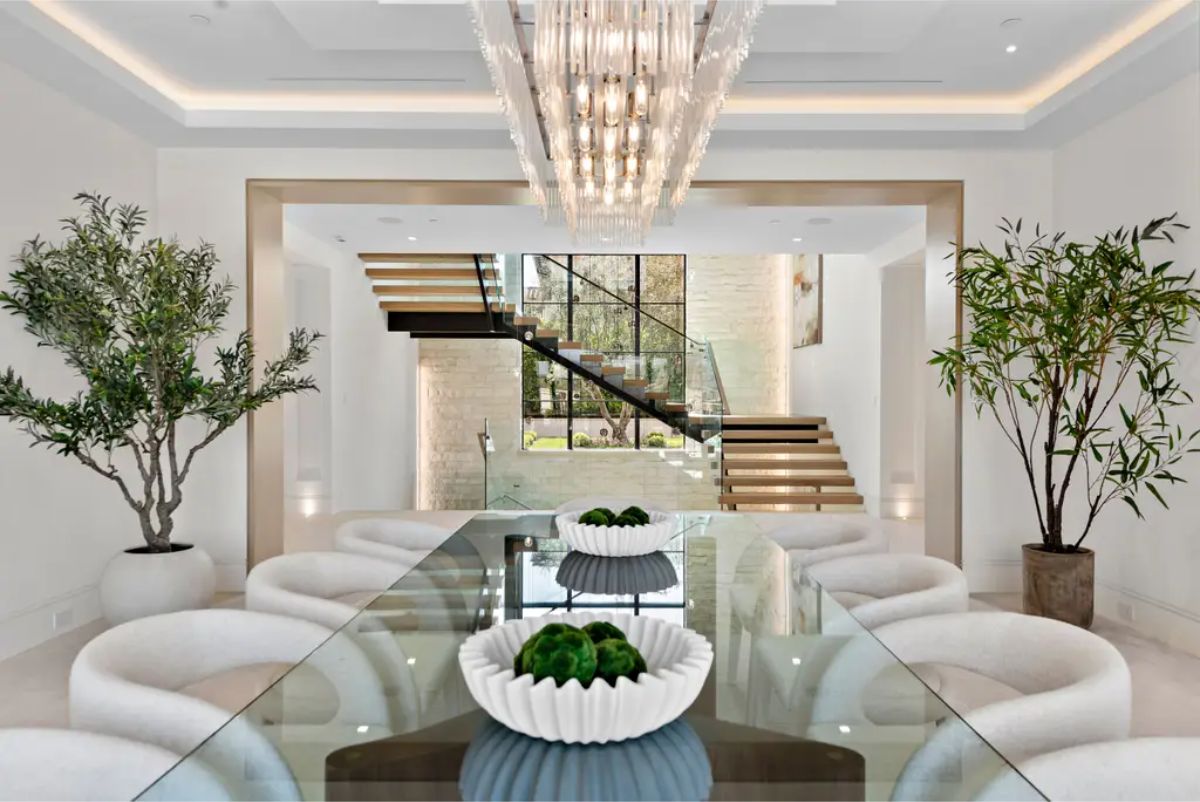 Glass dining table sits beneath a stunning chandelier, with a floating staircase adding architectural elegance. 