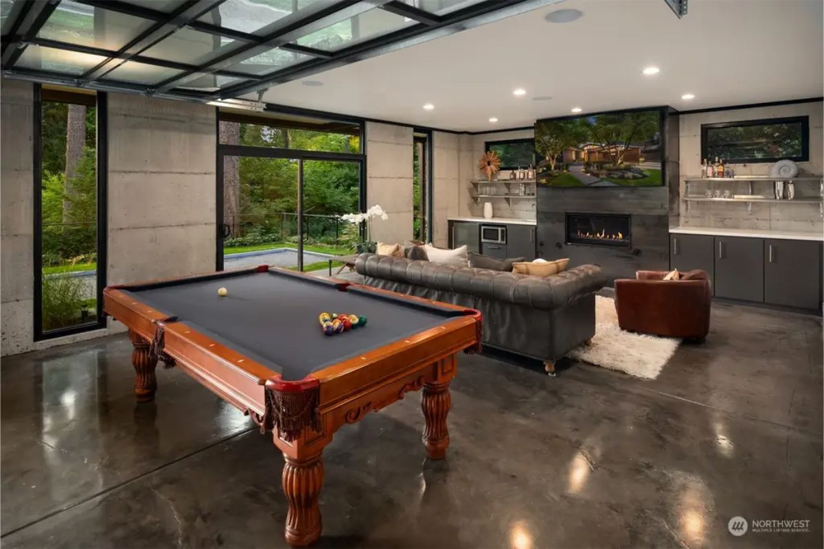 Game room and lounge with a classic wooden pool table, a fireplace, and large windows overlooking nature. 