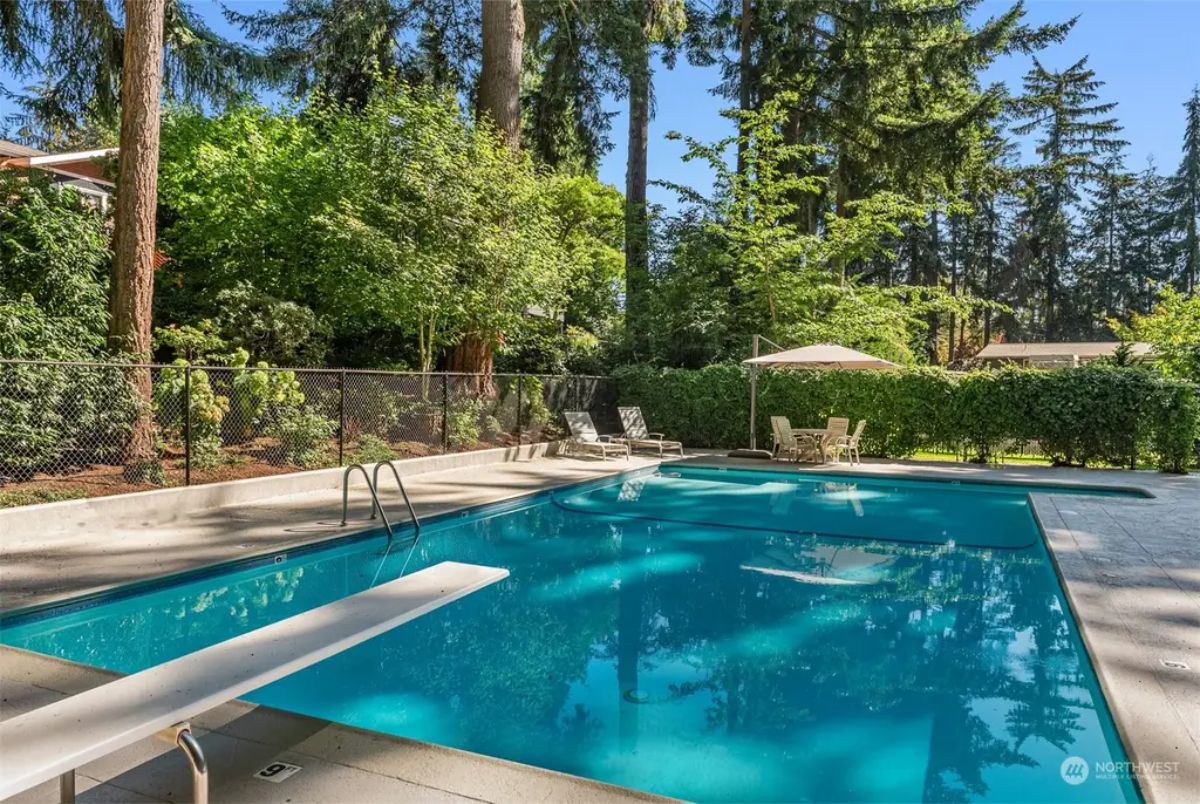 Outdoor swimming pool surrounded by lush greenery and tall trees, featuring a diving board and lounge chairs. 