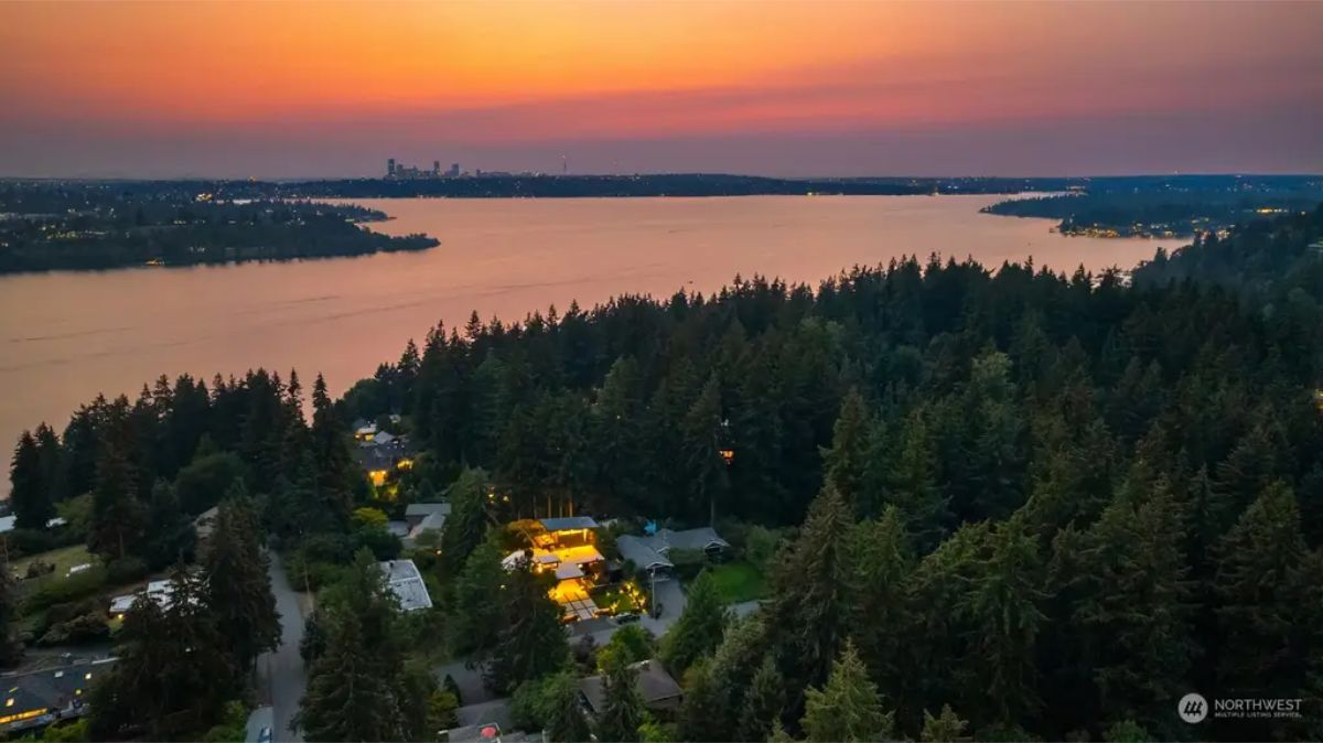 Aerial view of a lake at sunset, with a city skyline in the background and homes nestled among dense trees. 