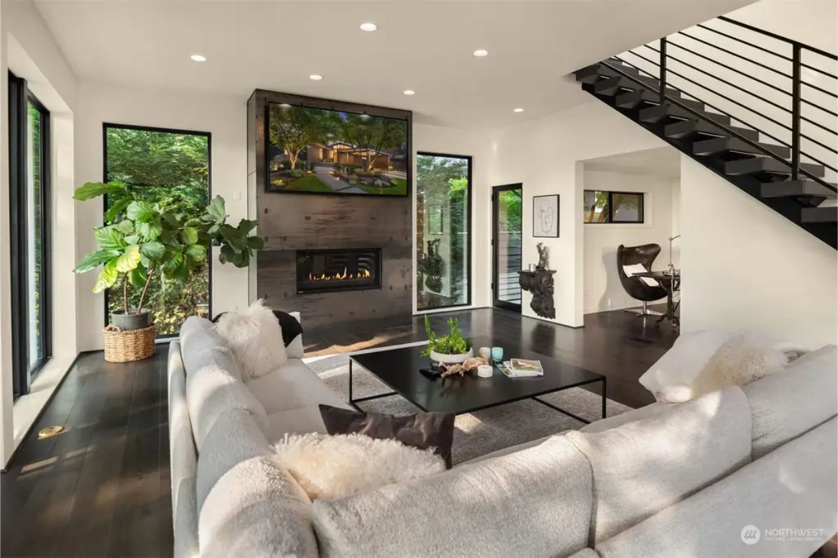 Open-concept living space with a fireplace, neutral-toned furniture, and a view of surrounding greenery. 