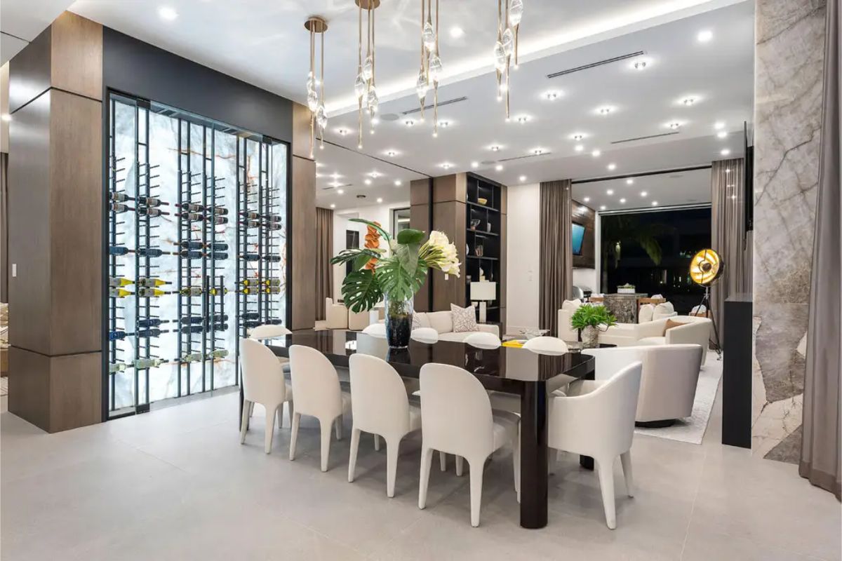 Contemporary dining space with a glossy dark wood table, white chairs, and a glass wine display accentuated by pendant lighting. 