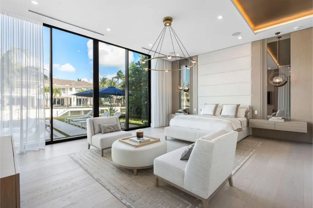 Bright master bedroom with floor-to-ceiling windows, soft neutral tones, and cozy seating facing a tranquil outdoor view. 