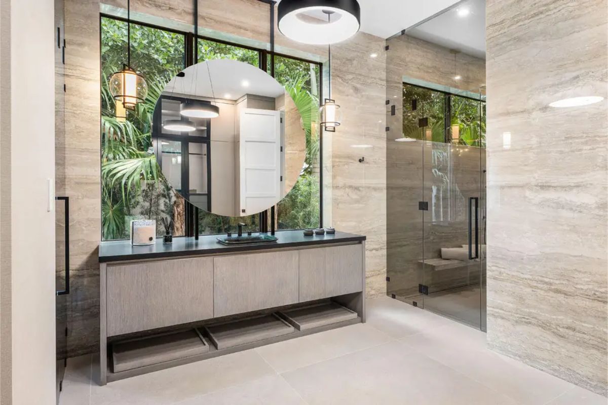 Contemporary bathroom showcasing a round mirror, dark countertop, and glass-enclosed shower with stone tile walls. 