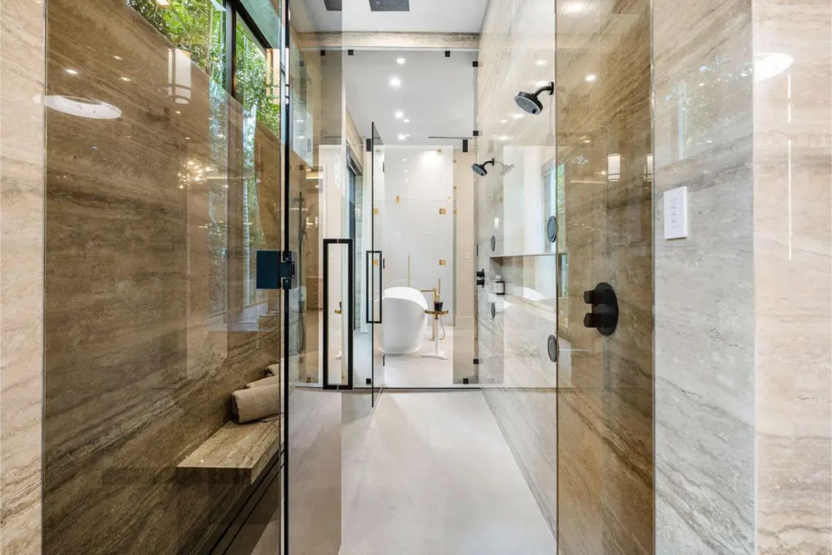 Luxurious bathroom with dual rainfall showerheads, glass enclosure, and a freestanding white bathtub in the background. 
