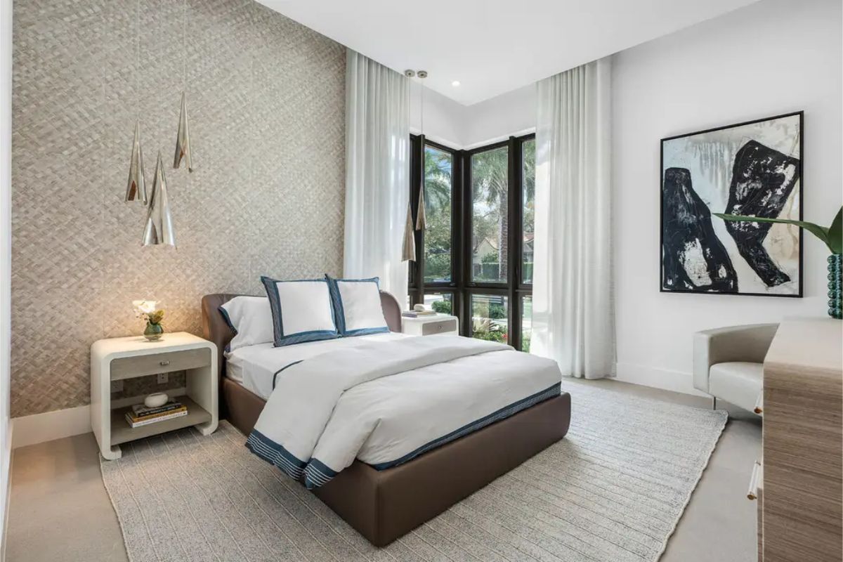 Bedroom featuring a cozy bed with blue-trimmed white linens, pendant lights, and a large window offering natural light and garden views. 