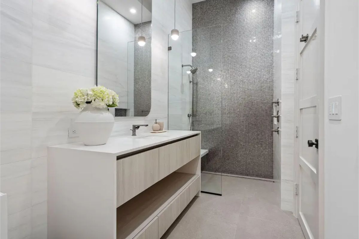 Minimalist bathroom design with a white vanity, large mirror, and a mosaic-tiled shower area. 