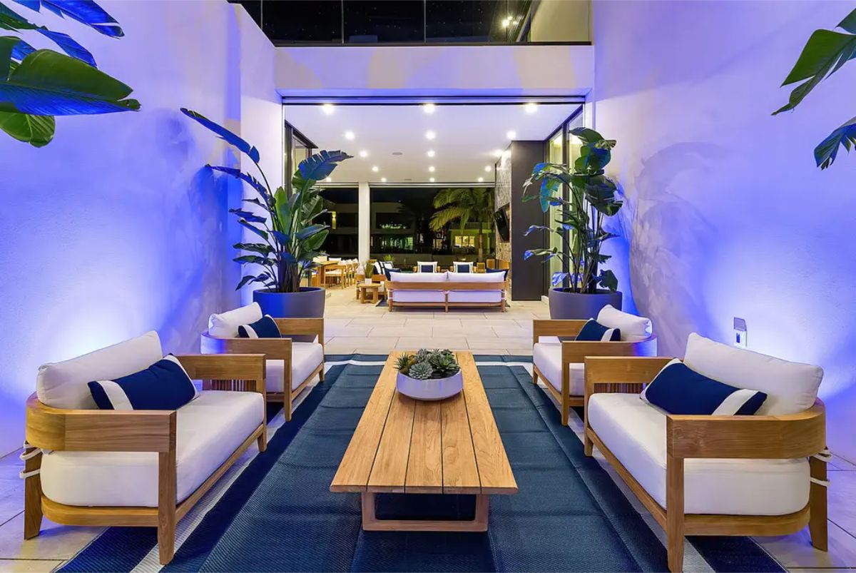 Cozy outdoor lounge with wooden furniture, white cushions, and blue lighting enhancing tropical plants. 