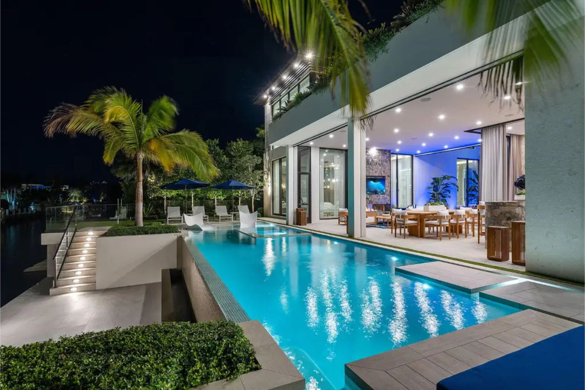 Illuminated pool stretches alongside an open-plan outdoor dining and lounge area under a modern, well-lit canopy. 