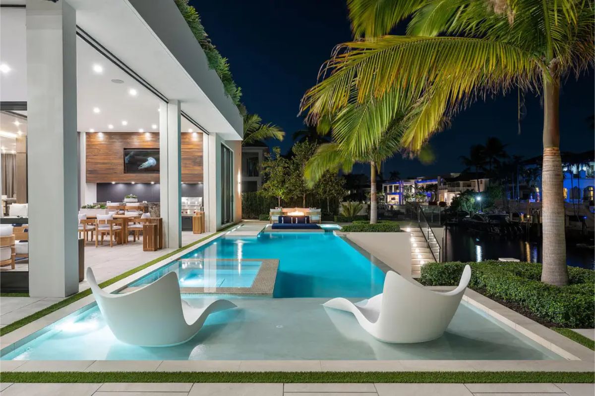 Sleek poolside lounge chairs sit in a shallow water area next to a luxurious swimming pool with palm trees and ambient night lighting. 