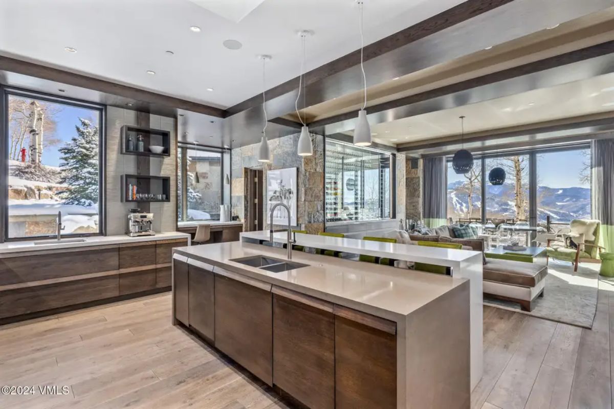 A sleek kitchen with minimalist cabinetry and dual islands overlooks a bright living area with panoramic mountain scenery.