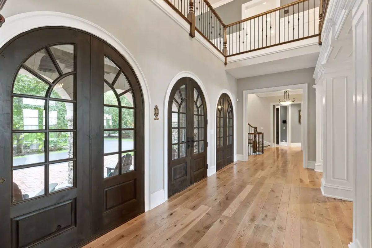 Bright entryway showcases wide-plank hardwood flooring and a series of dark wood arched double doors with glass panels.