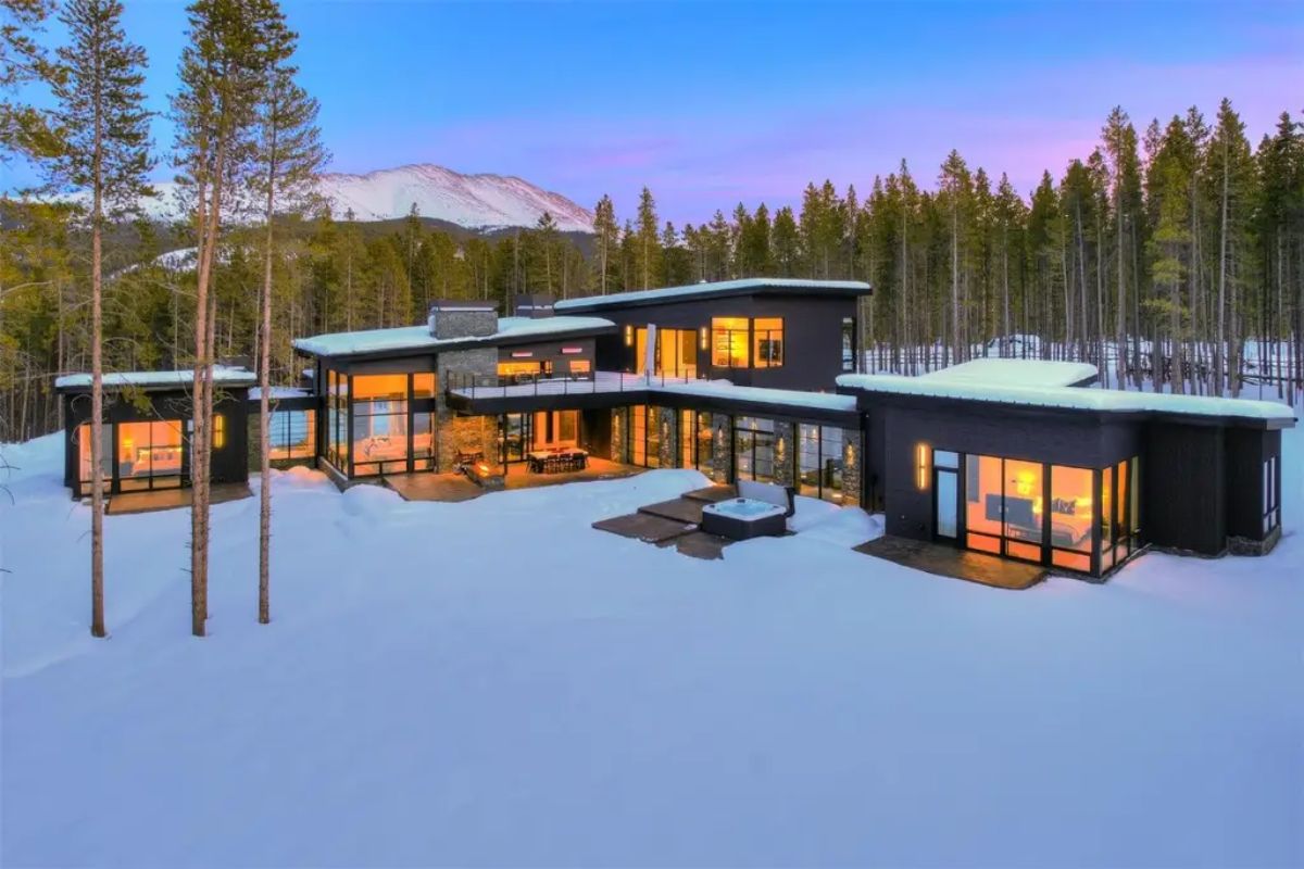 Modern mountain home is surrounded by snow and tall trees, with warm interior lighting visible through large glass windows.