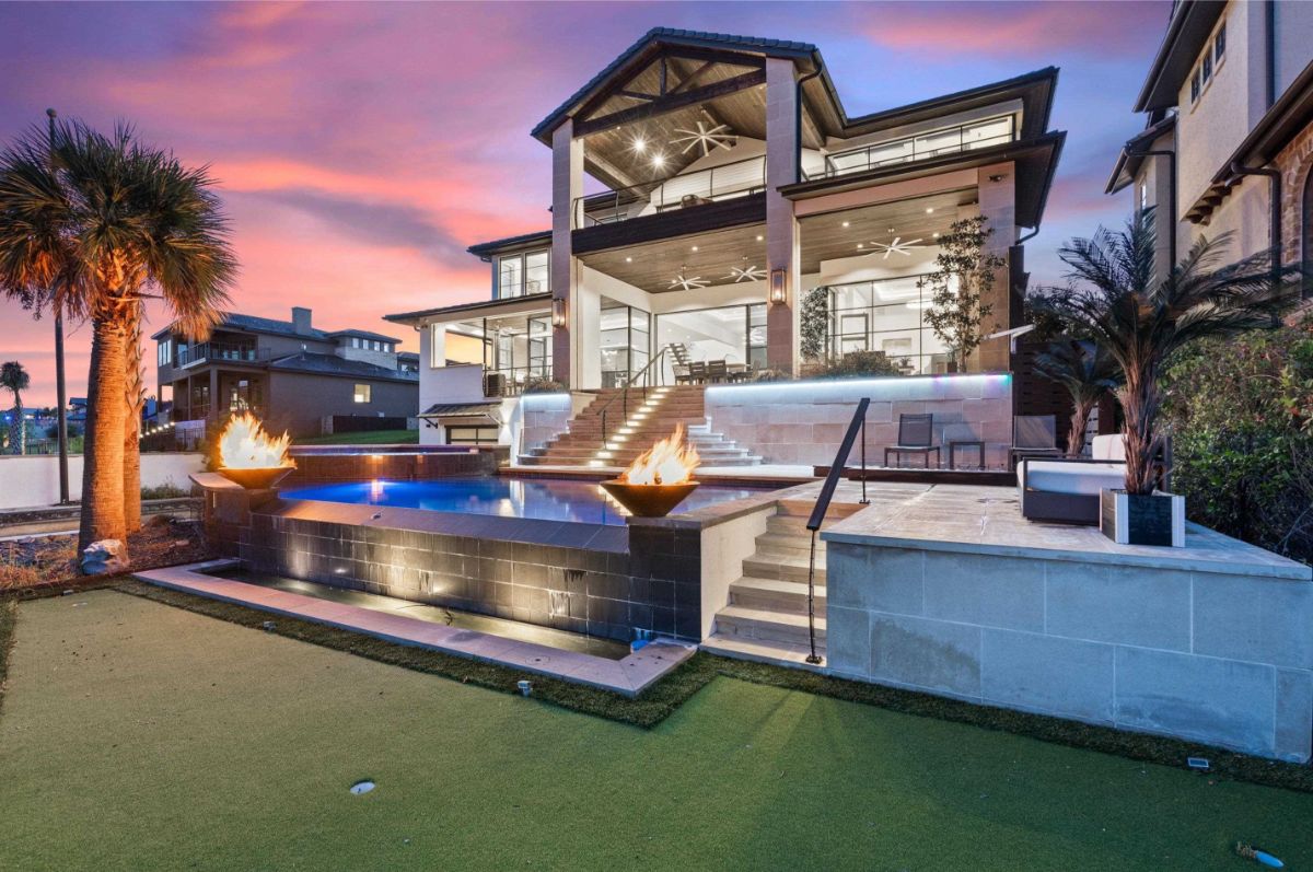 Stunning outdoor pool area features a sleek modern design with fire bowls, cascading steps, and ambient lighting against a colorful sunset sky.