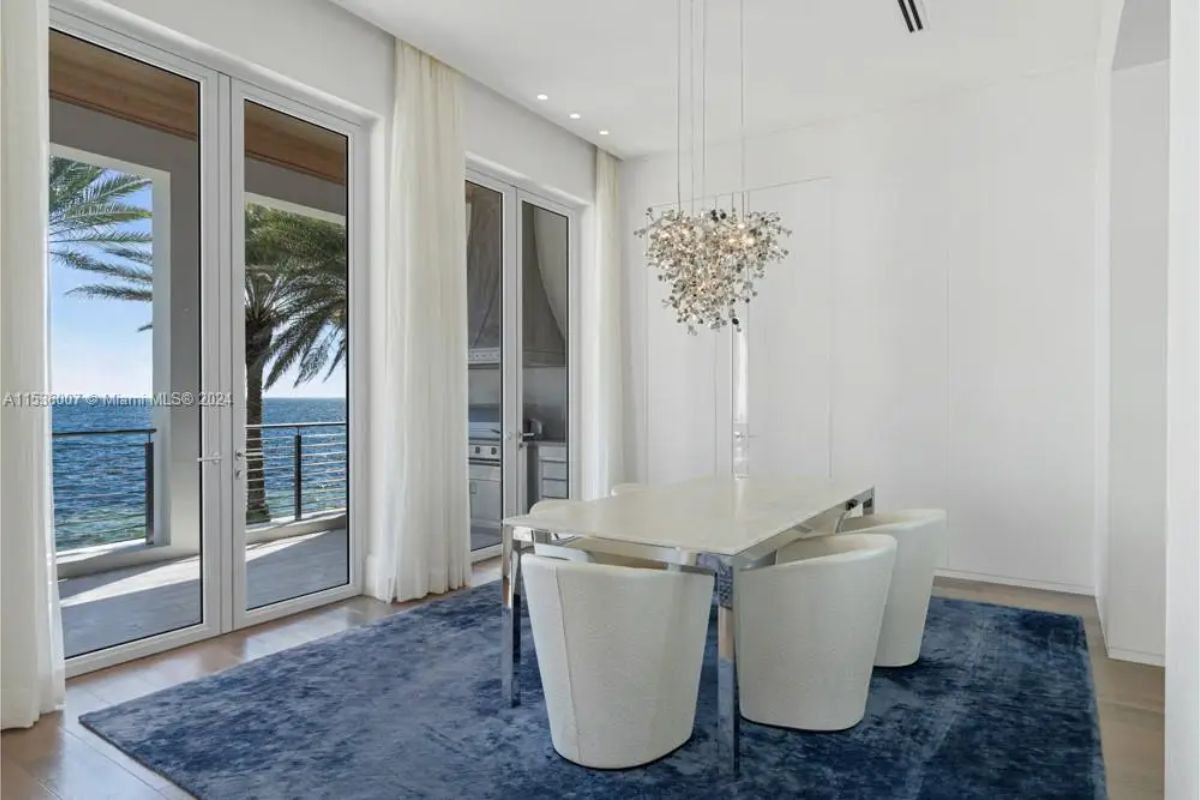 Minimalistic dining area showcasing a white table, plush white chairs, a unique cascading pendant light, and floor-to-ceiling windows overlooking the ocean. 