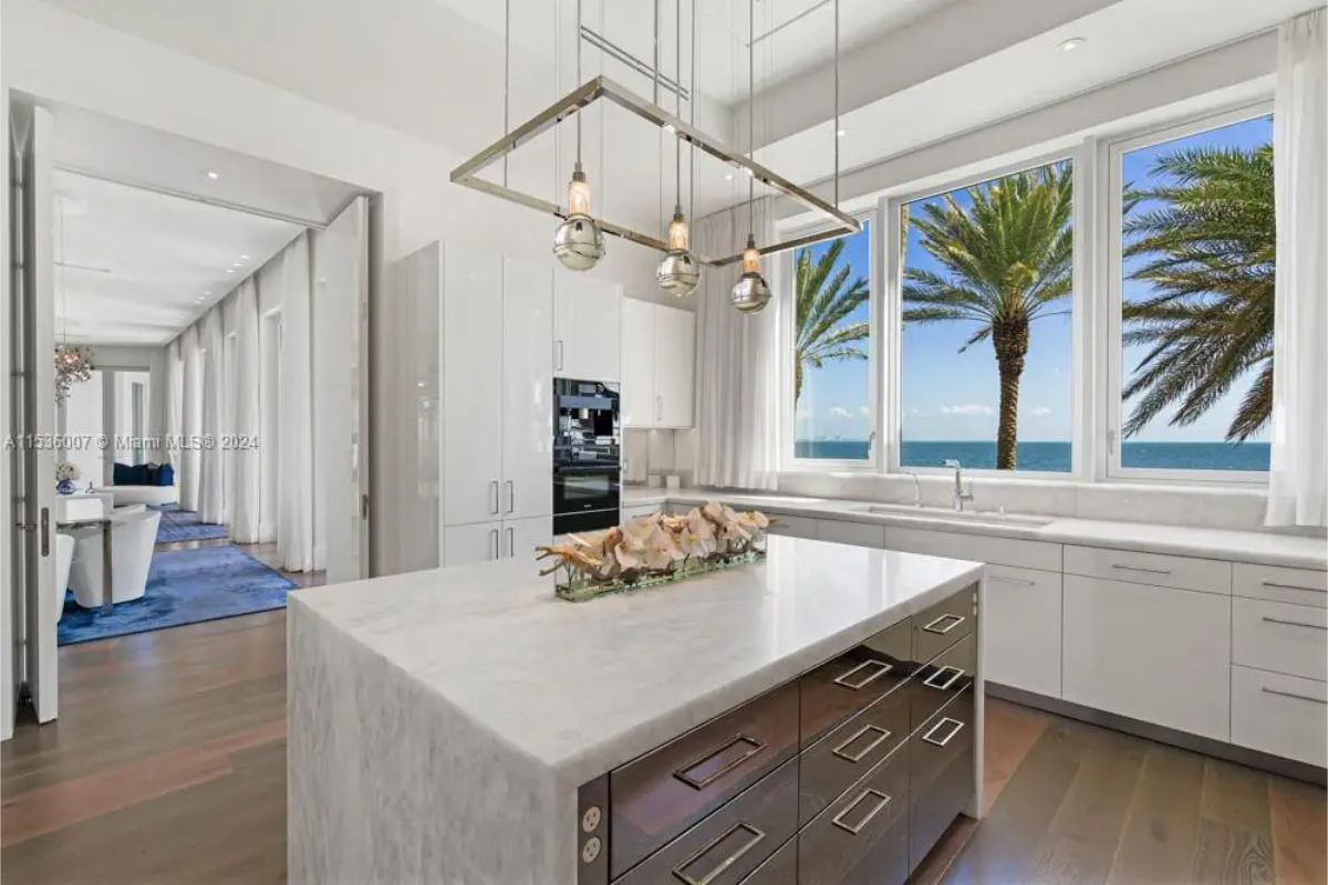 Bright kitchen with a marble island, sleek white cabinetry, industrial-style pendant lights, and large windows framing picturesque ocean and palm tree views. 