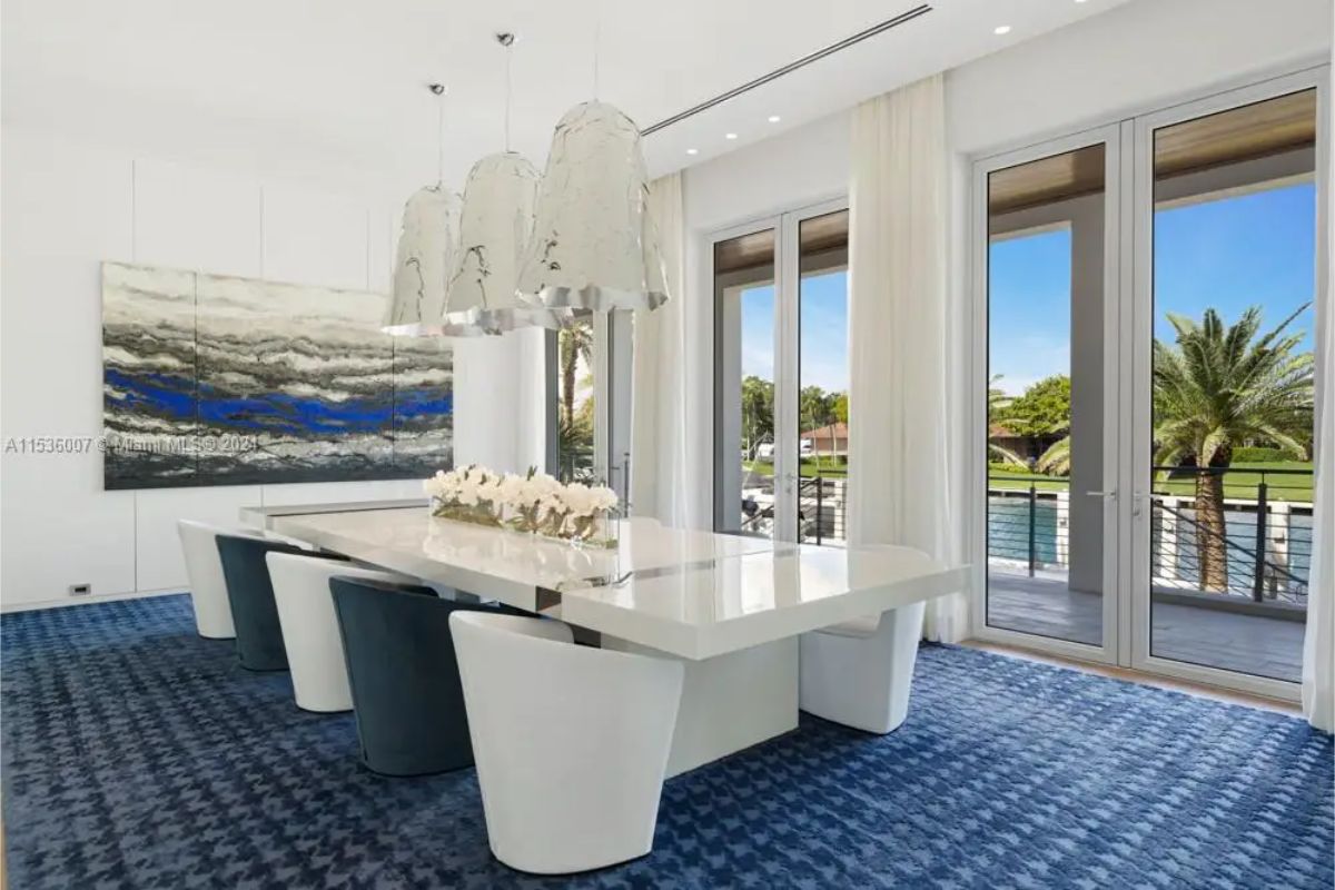 Elegant dining room with a glossy white table, a mix of white and dark blue chairs, and large glass doors offering views of palm trees and the waterfront. 