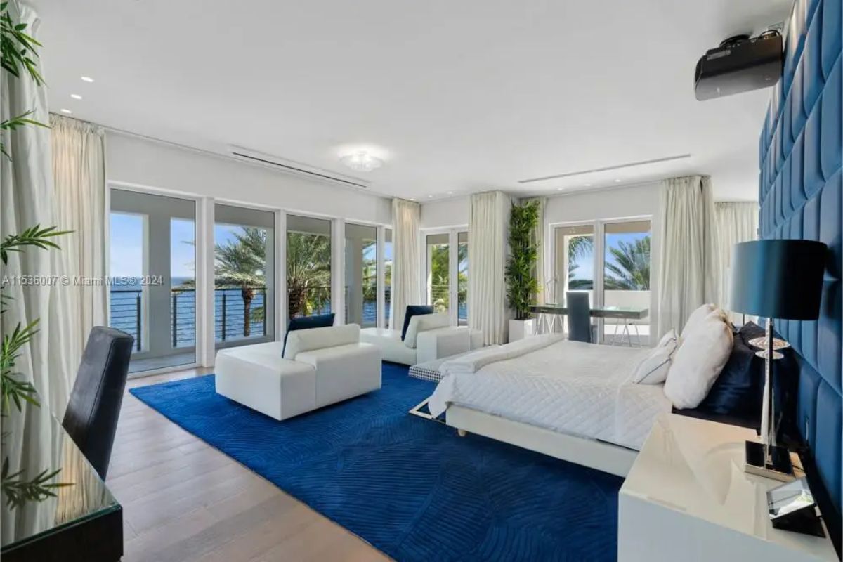 Spacious bedroom highlighted by a deep blue carpet, modern white furniture, floor-to-ceiling windows, and panoramic views of the surrounding water and palm trees. 