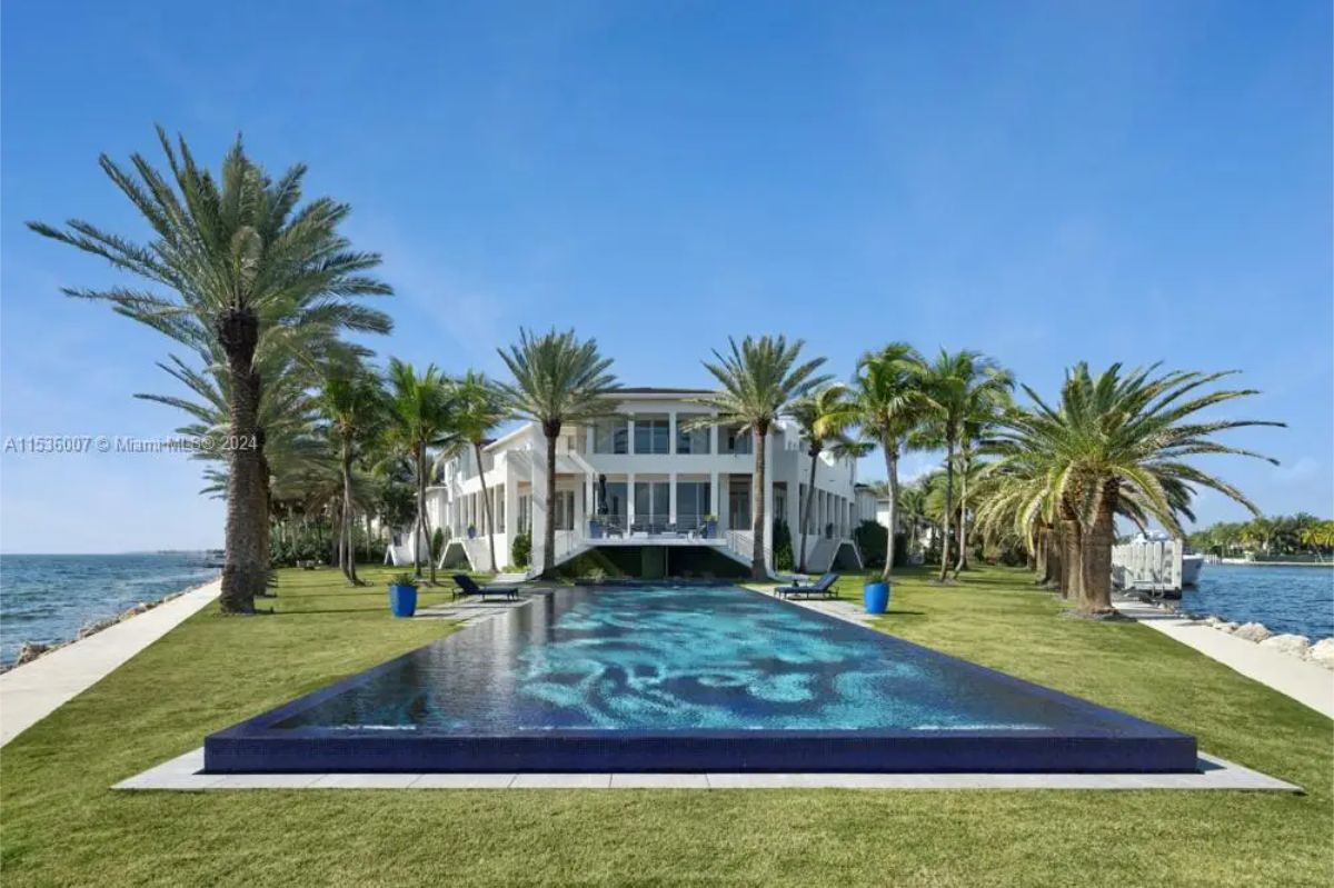 Daytime scene highlights a waterfront house with a rectangular infinity pool, surrounded by palm trees and neatly trimmed grass. 
