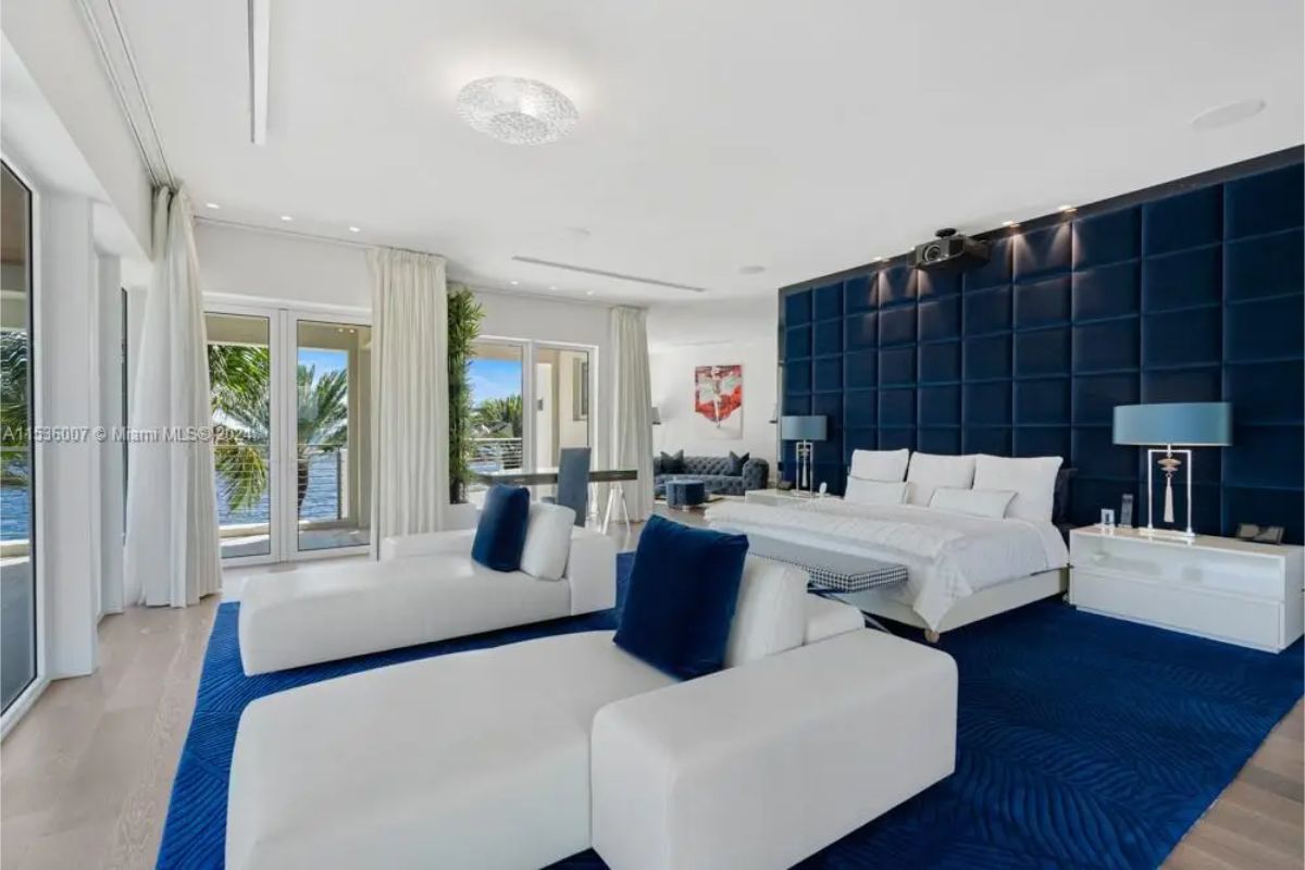 Bedroom features a white bed with blue accents, two white lounge chairs, and floor-to-ceiling windows overlooking the water. 