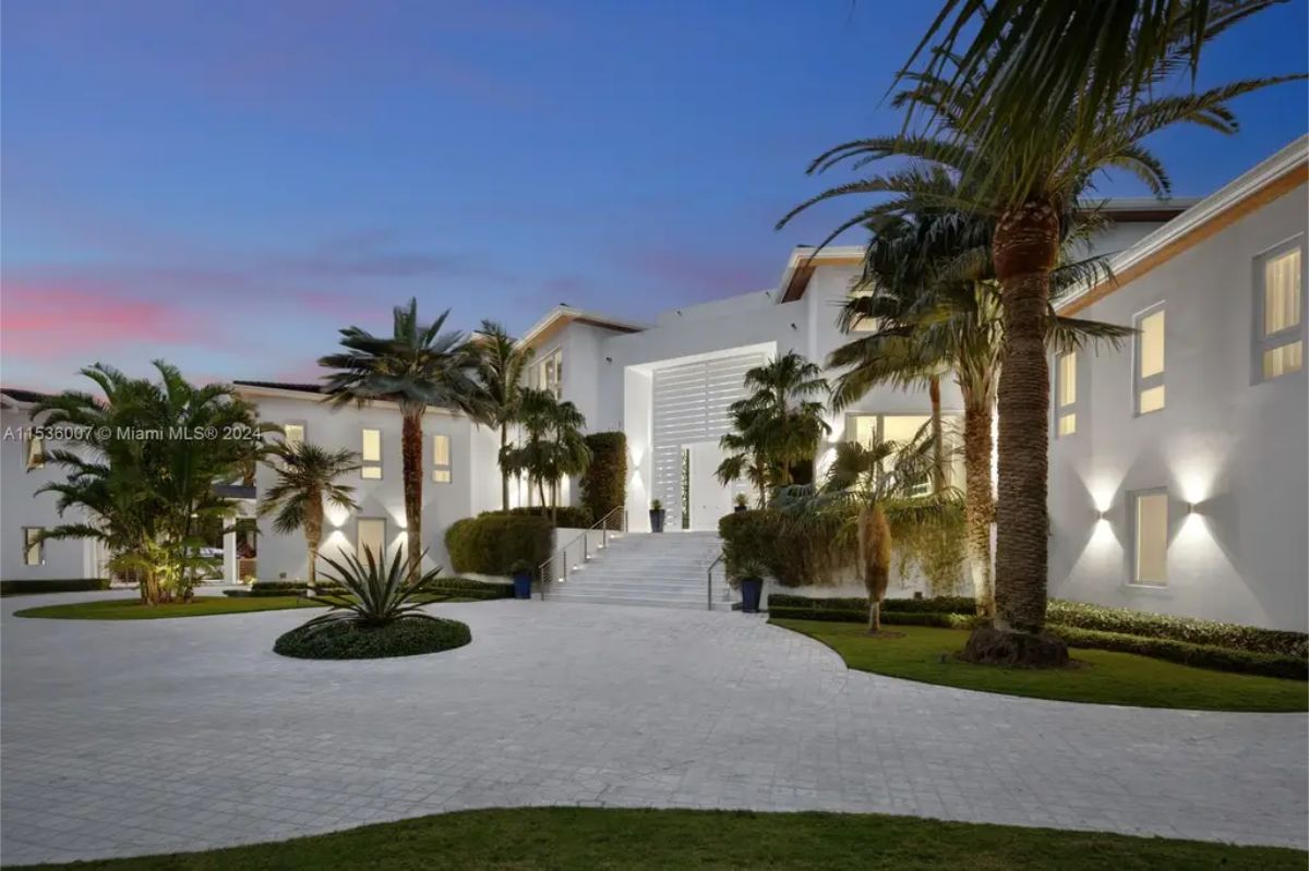 Elegant driveway leading to a contemporary mansion with ambient lighting, tropical landscaping, and a dramatic sunset sky. 