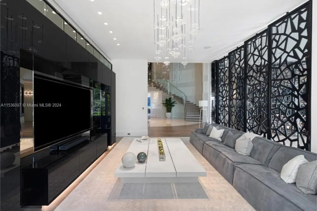 Modern entertainment room featuring a sleek black TV wall, long gray sectional sofa, and artistic geometric black partitions with hanging glass light fixtures. 
