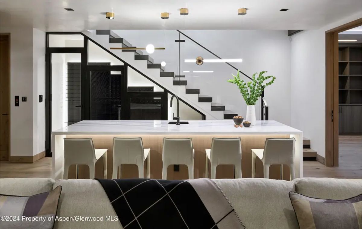 Sleek kitchen design with a marble island, barstools, and modern lighting under a glass-railed staircase. 