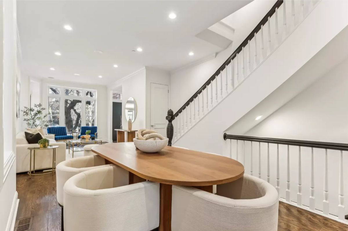 A wooden dining table with rounded-edge chairs sits near a modern staircase with white railings and dark handrails.