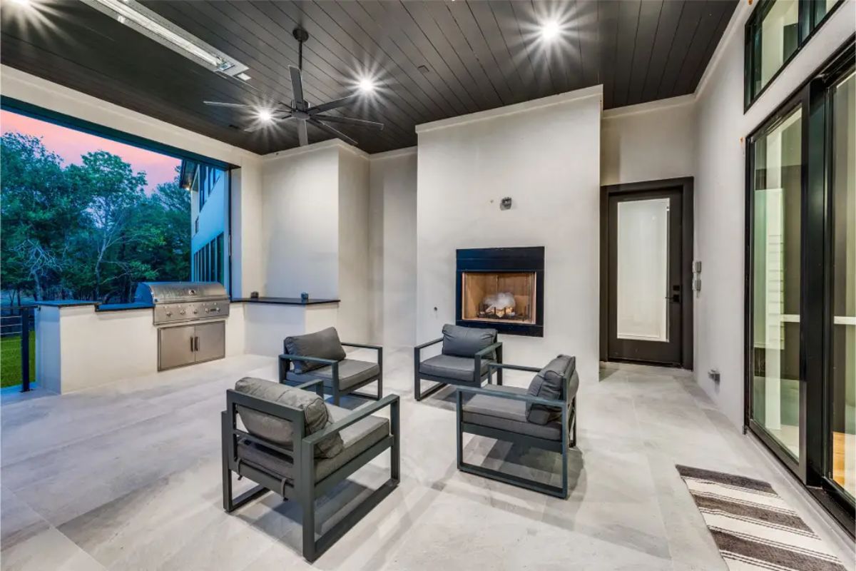 Outdoor patio features a built-in grill, fireplace, and comfortable lounge chairs under a black ceiling. 