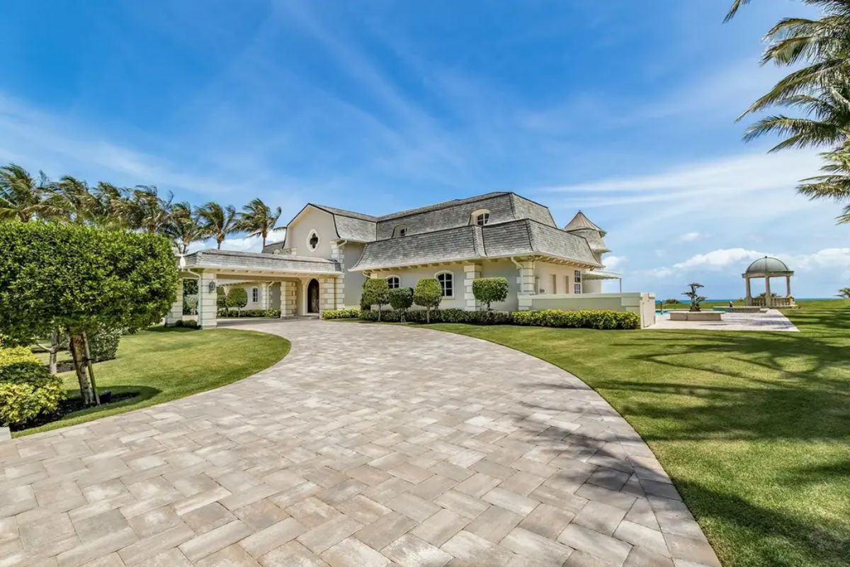Circular driveway curves gracefully toward the main entrance, surrounded by lush greenery and a view of the ocean beyond. 