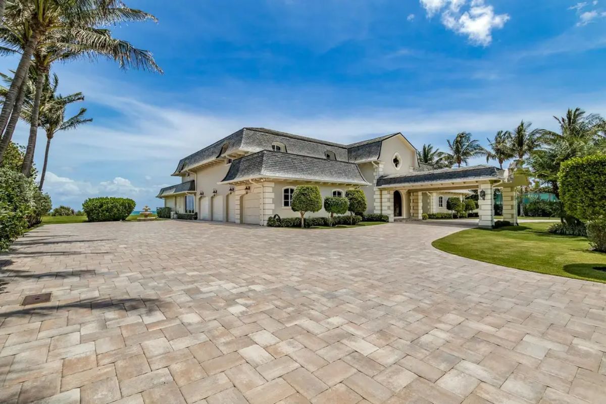 Spacious paved driveway leads to a luxurious, light-colored mansion with elegant landscaping and palm trees. 