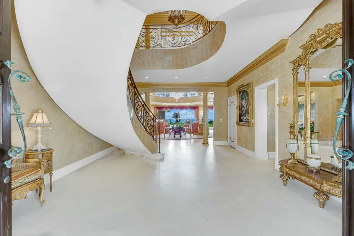 Bright entry hall opens into an opulent living area, with a dramatic staircase serving as a focal point under a detailed ceiling. 