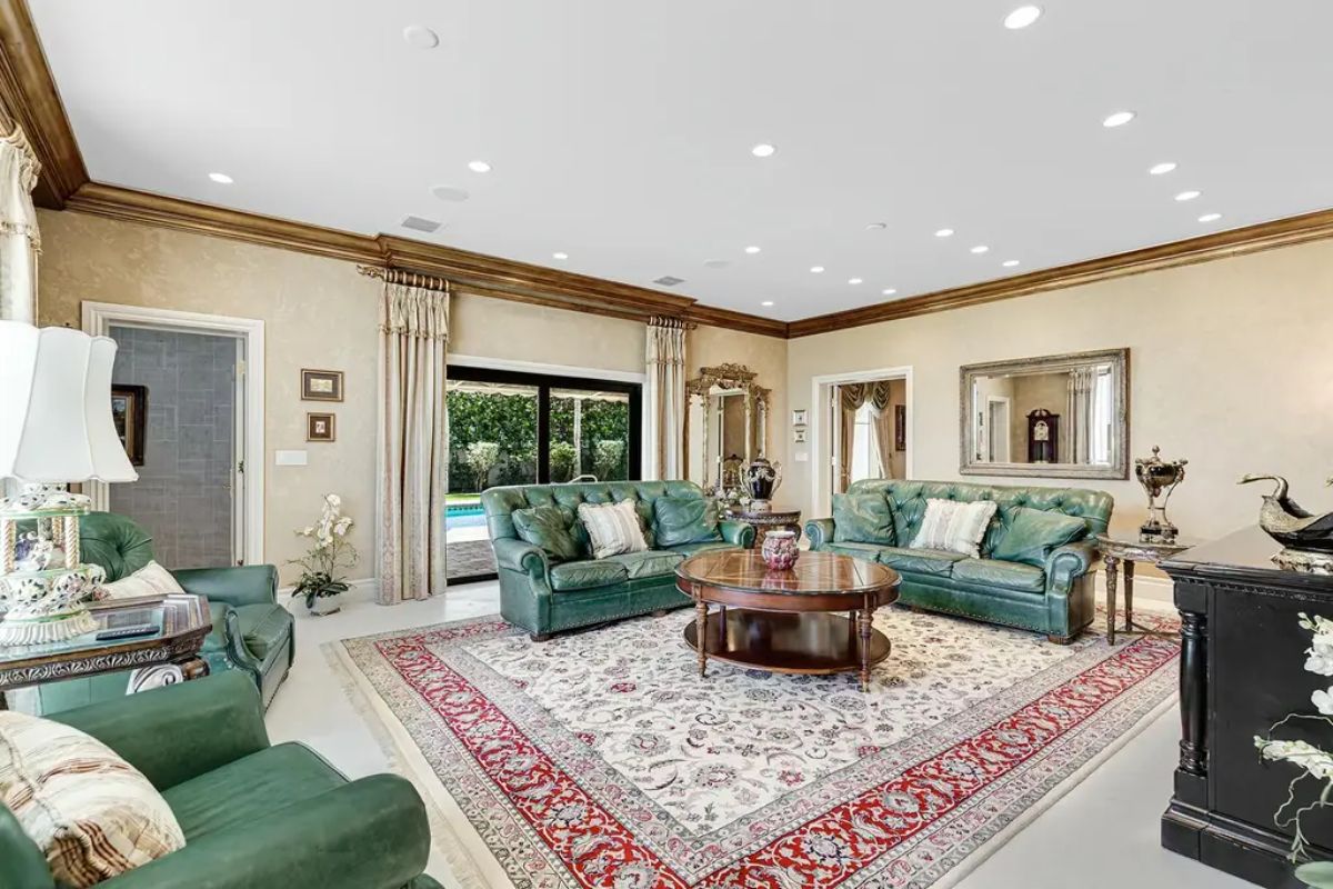 Elegant living room features green leather sofas, a large ornate rug, and sliding glass doors that open to a poolside view. 