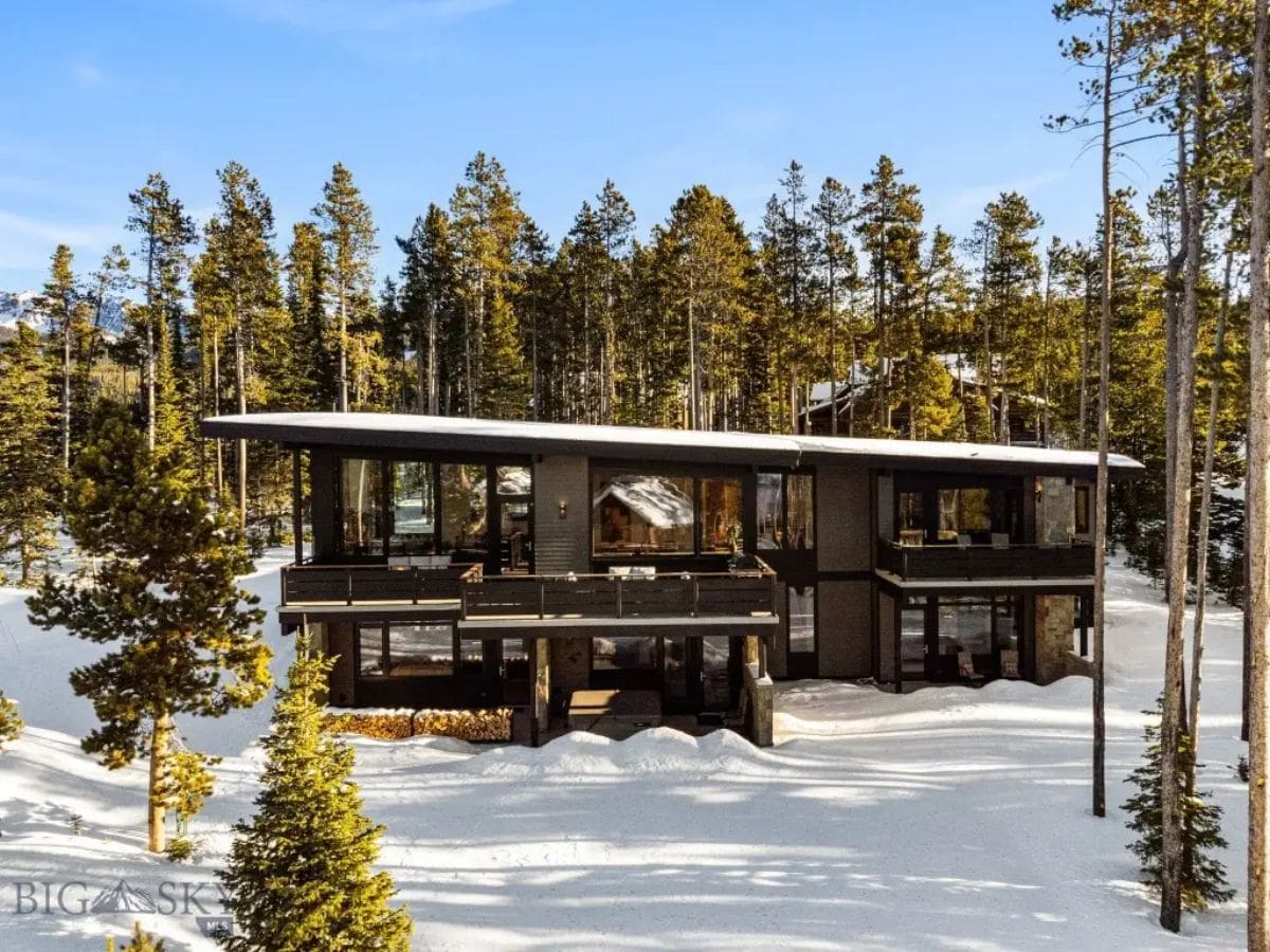 A contemporary two-story house with dark exterior finishes is set among tall pine trees in a snowy landscape.