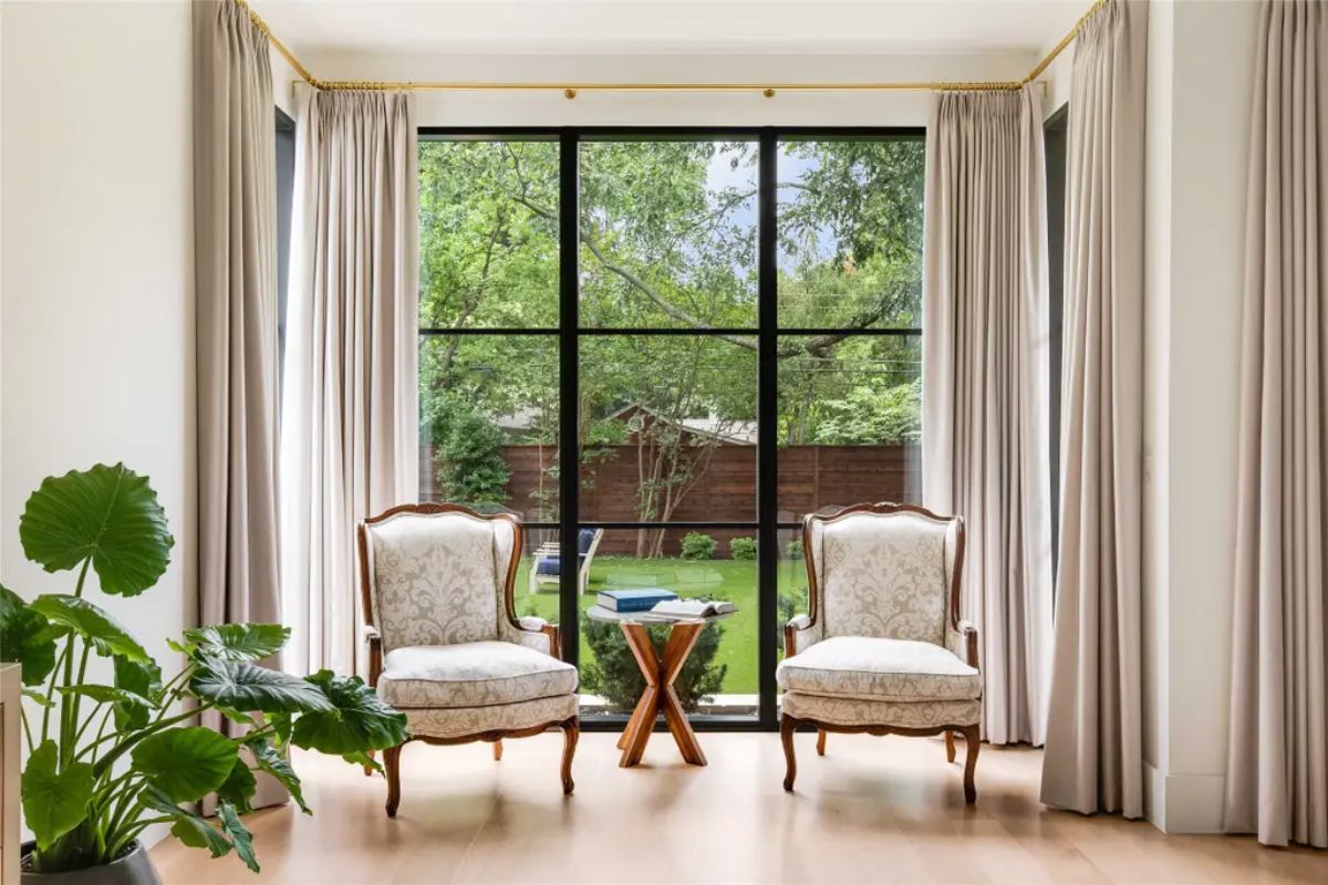 Elegant sitting area with two antique-style chairs, a wooden side table, and large windows overlooking a lush backyard. 