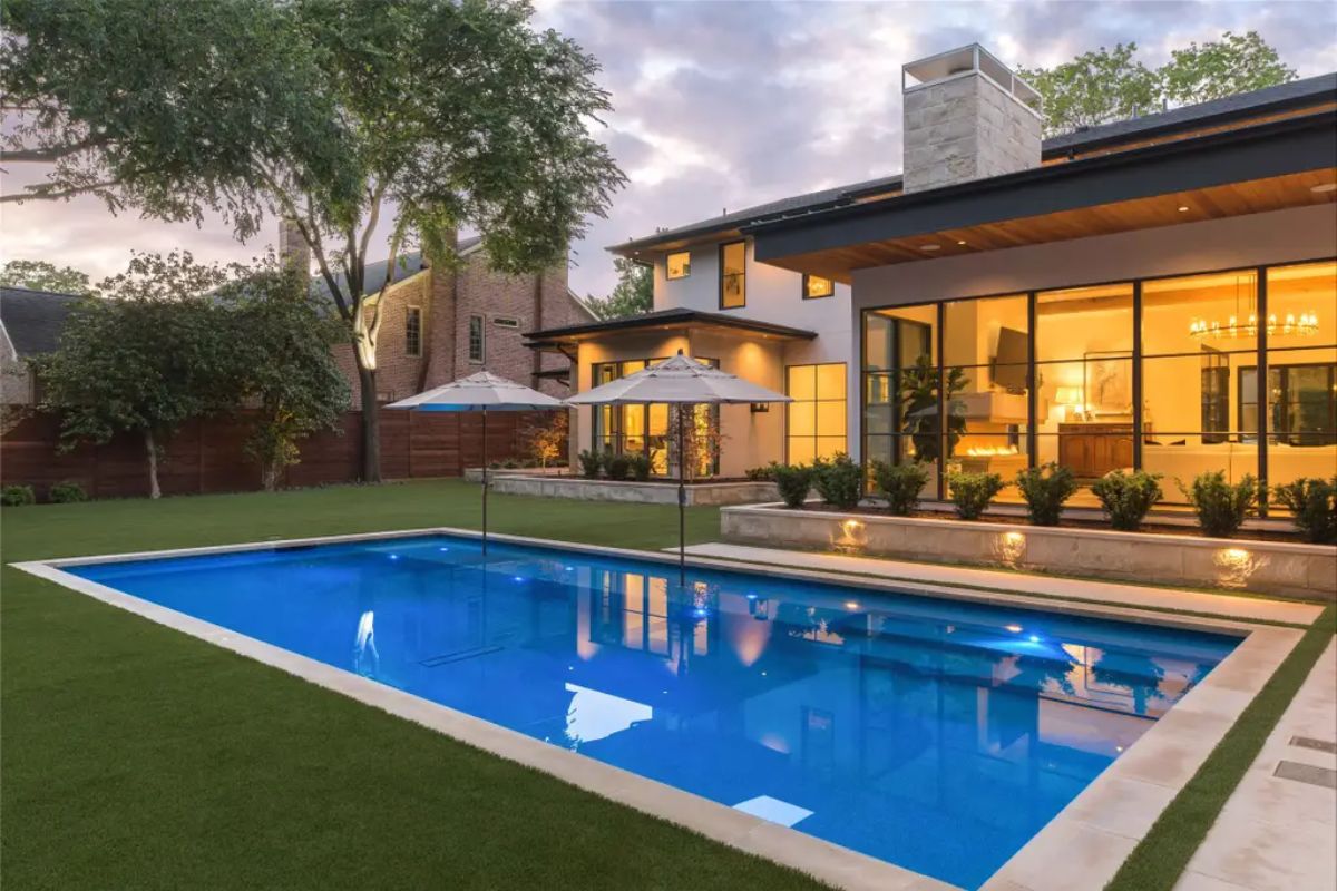 Evening view of the backyard pool area with ambient lighting, lush landscaping, and stylish umbrellas. 