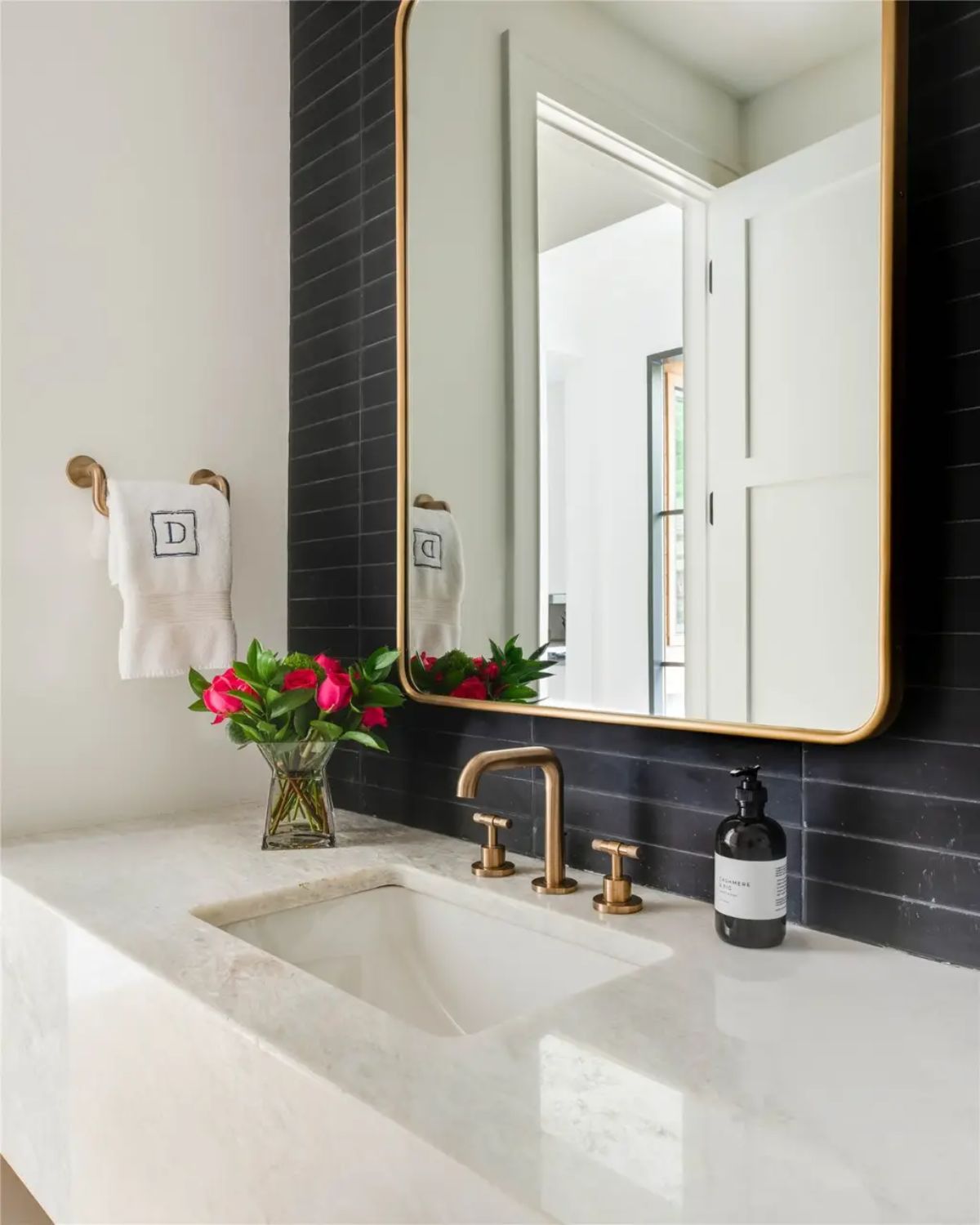 Stylish bathroom vanity featuring a black tile accent wall, gold-framed mirror, and marble countertop. 