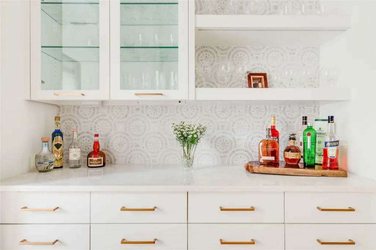Stylish home bar with a white and gold color scheme, glass shelving, and a decorative backsplash. 