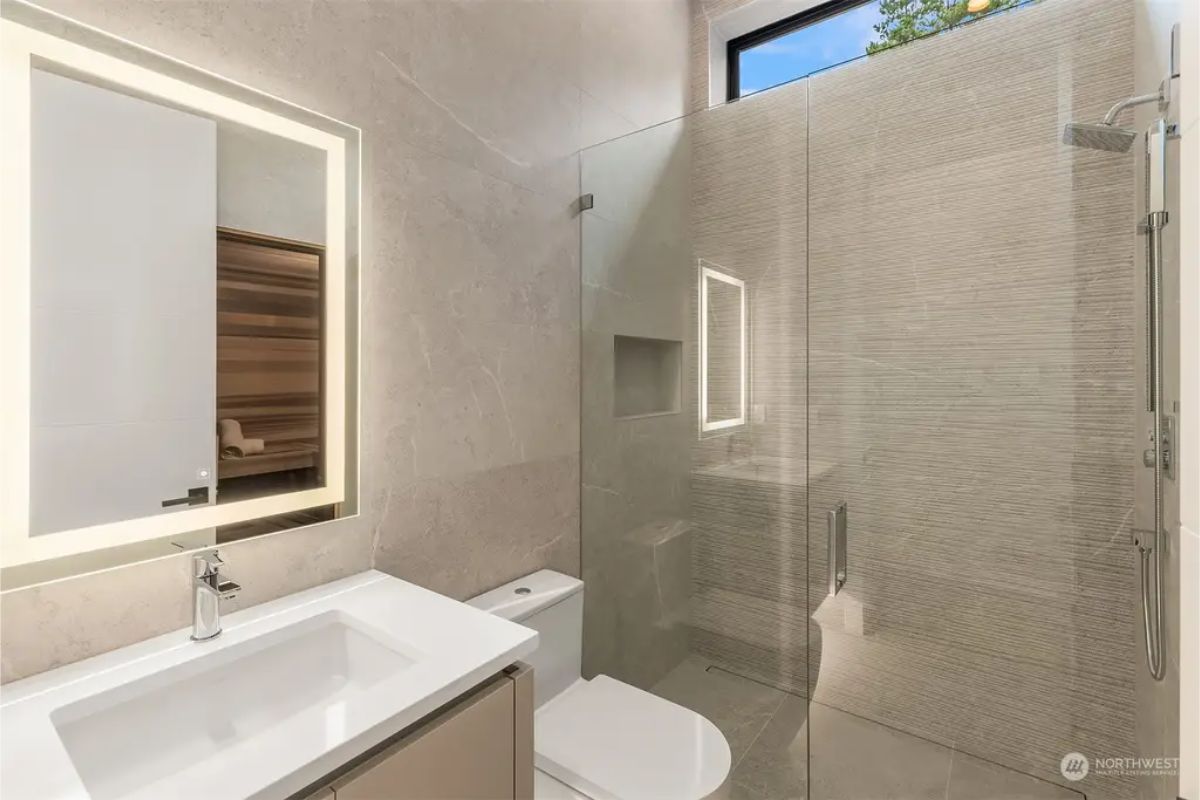 Compact modern bathroom with a glass-enclosed shower, illuminated mirror, and subtle textures. 