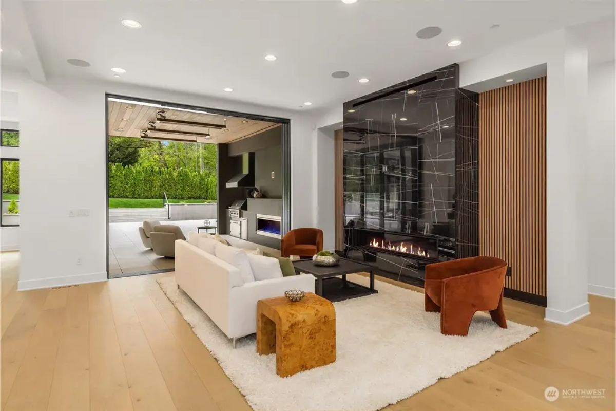 Spacious living room with a modern fireplace, wood-paneled wall, and sliding doors leading to an outdoor lounge and kitchen. 