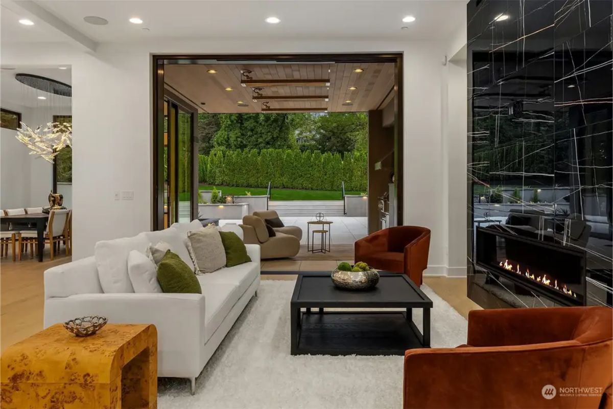 Cozy seating area with white and brown furniture, a black marble fireplace, and large glass doors opening to a manicured backyard. 