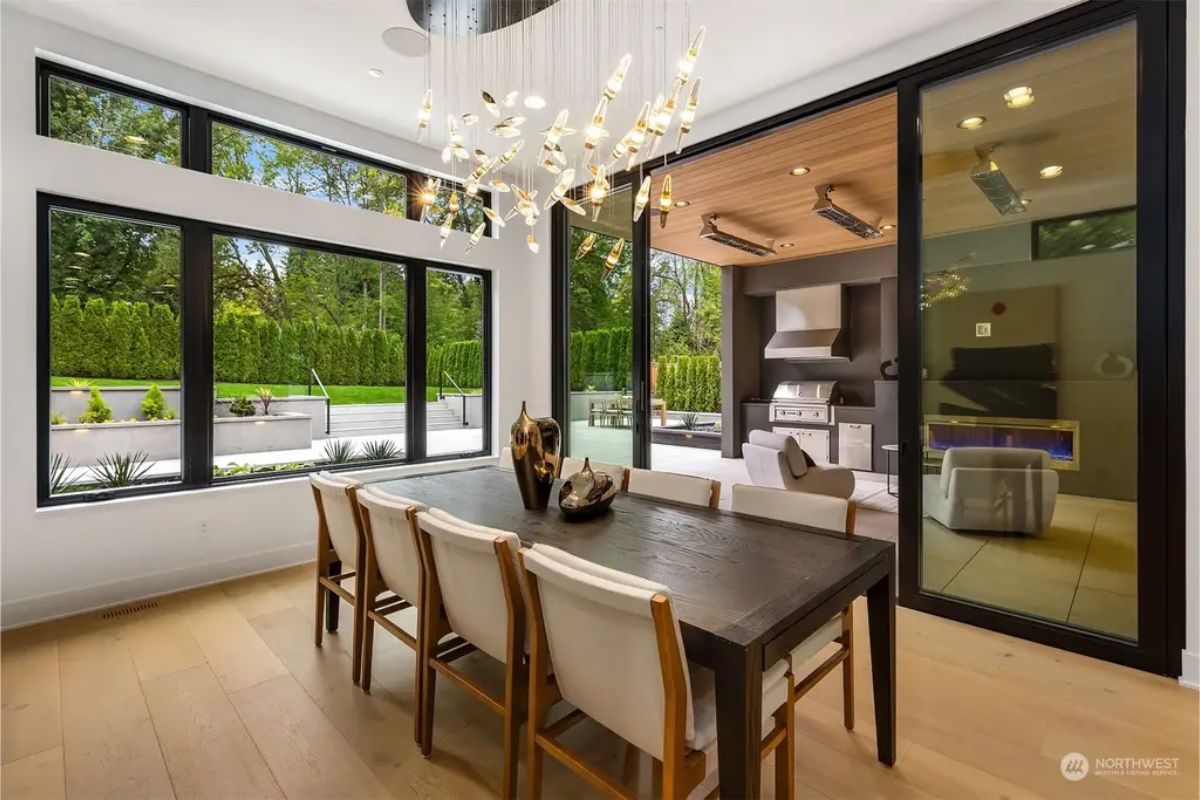Bright dining area with floor-to-ceiling windows, gold-accented chandelier, and a view of the outdoor kitchen. 