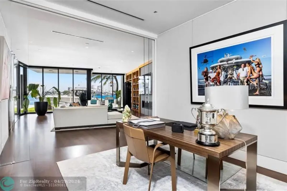 Home office setup with a wooden desk, leather chair, large framed artwork, and a view of a living area with expansive windows. 