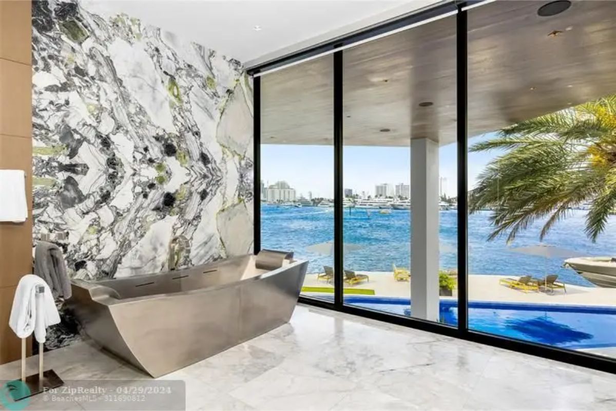 Freestanding metallic bathtub positioned next to floor-to-ceiling windows with a waterfront view and a marble accent wall. 