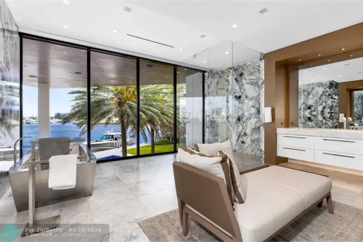 Spacious bathroom featuring a lounge chair, glass-enclosed shower, double sink vanity, and large windows overlooking water and palm trees. 
