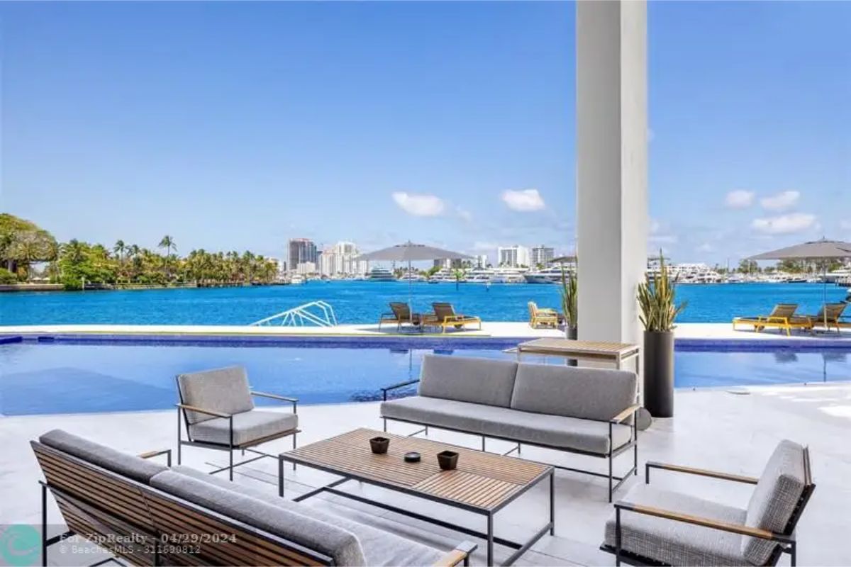 Poolside seating area with gray cushioned chairs, wooden tables, and views of the pool and waterfront city skyline. 