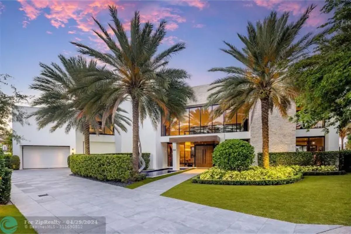 Contemporary house exterior featuring tall palm trees, manicured landscaping, and large glass windows reflecting the evening sky. 