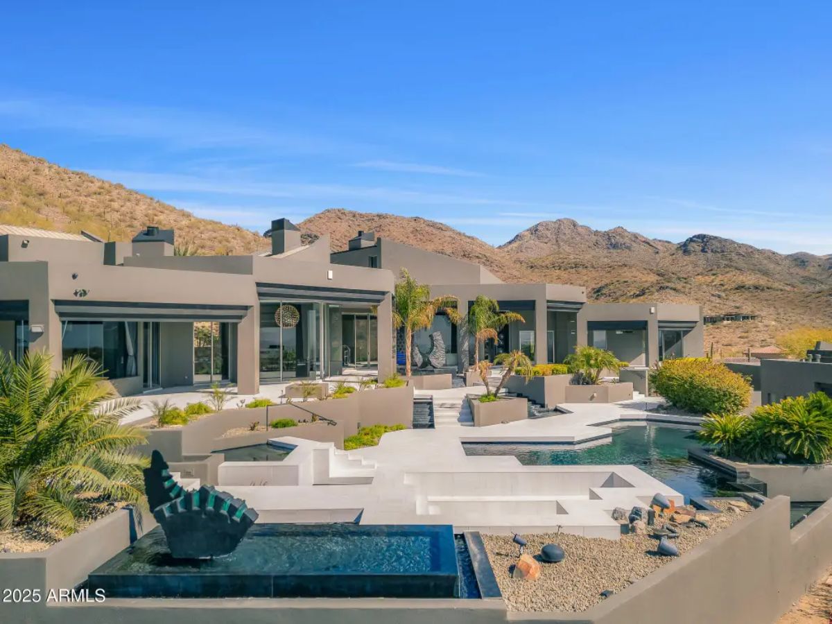 Luxurious modern home sits in a desert landscape, featuring an expansive outdoor space with pools, lounge areas, and lush greenery.